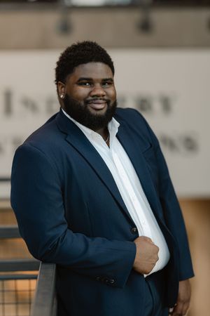 A smiling Black man in a navy blue suit and white shirt leans against a railing indoors.