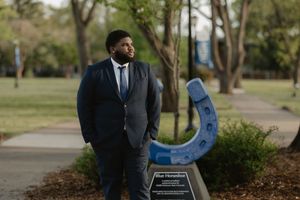 Young Black man in navy suit poses confidently outdoors near a blue horseshoe sculpture on a college campus.