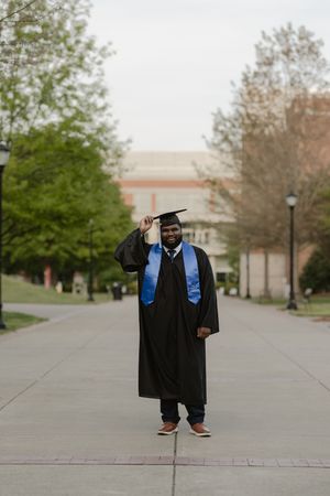 Smiling graduate in black cap and gown with blue stole poses on campus walkway surrounded by trees.
