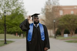 Graduate in black cap and gown with blue stole poses proudly on college campus walkway.