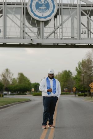 Graduate in white shirt and blue stole stands on road beneath university seal bridge outdoors.