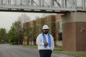 Graduate in cap and gown with hard hat stands on urban street near bridge, celebrating engineering achievement.
