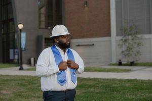 Man in white hard hat and blue graduation stole stands outdoors near brick building on campus.