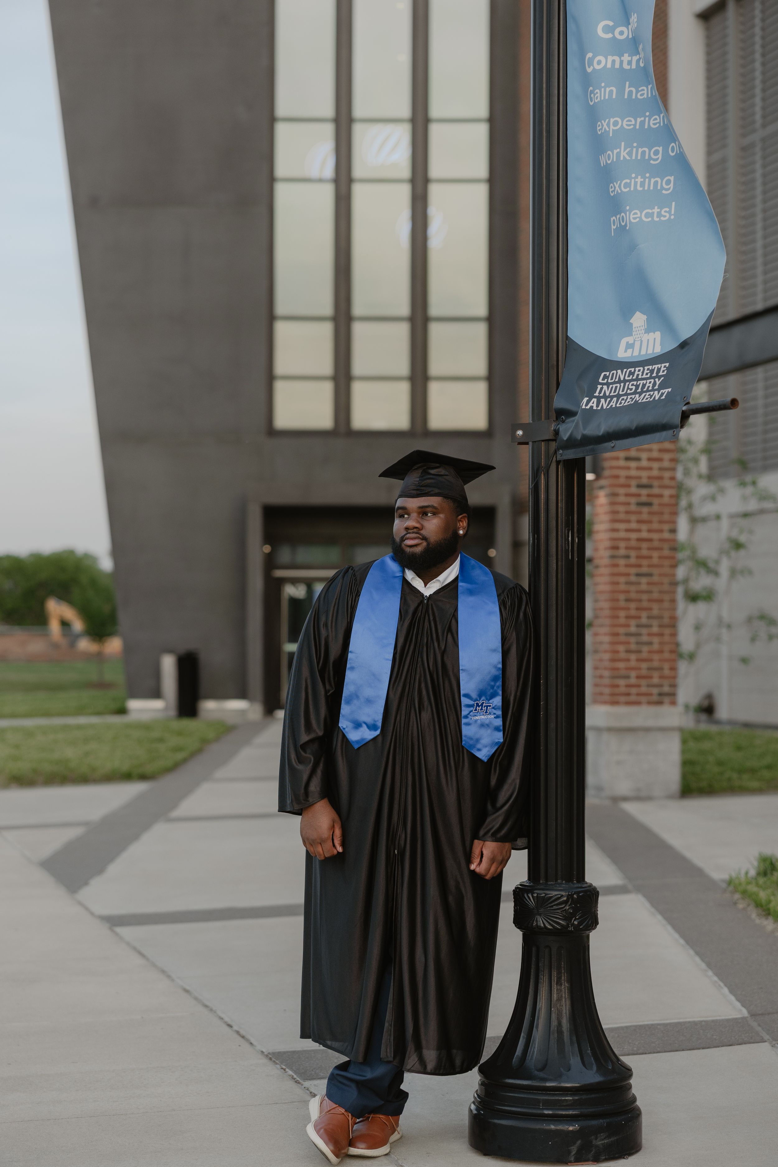 Graduate in black cap and gown with blue stole poses by a lamp post outside a university building.