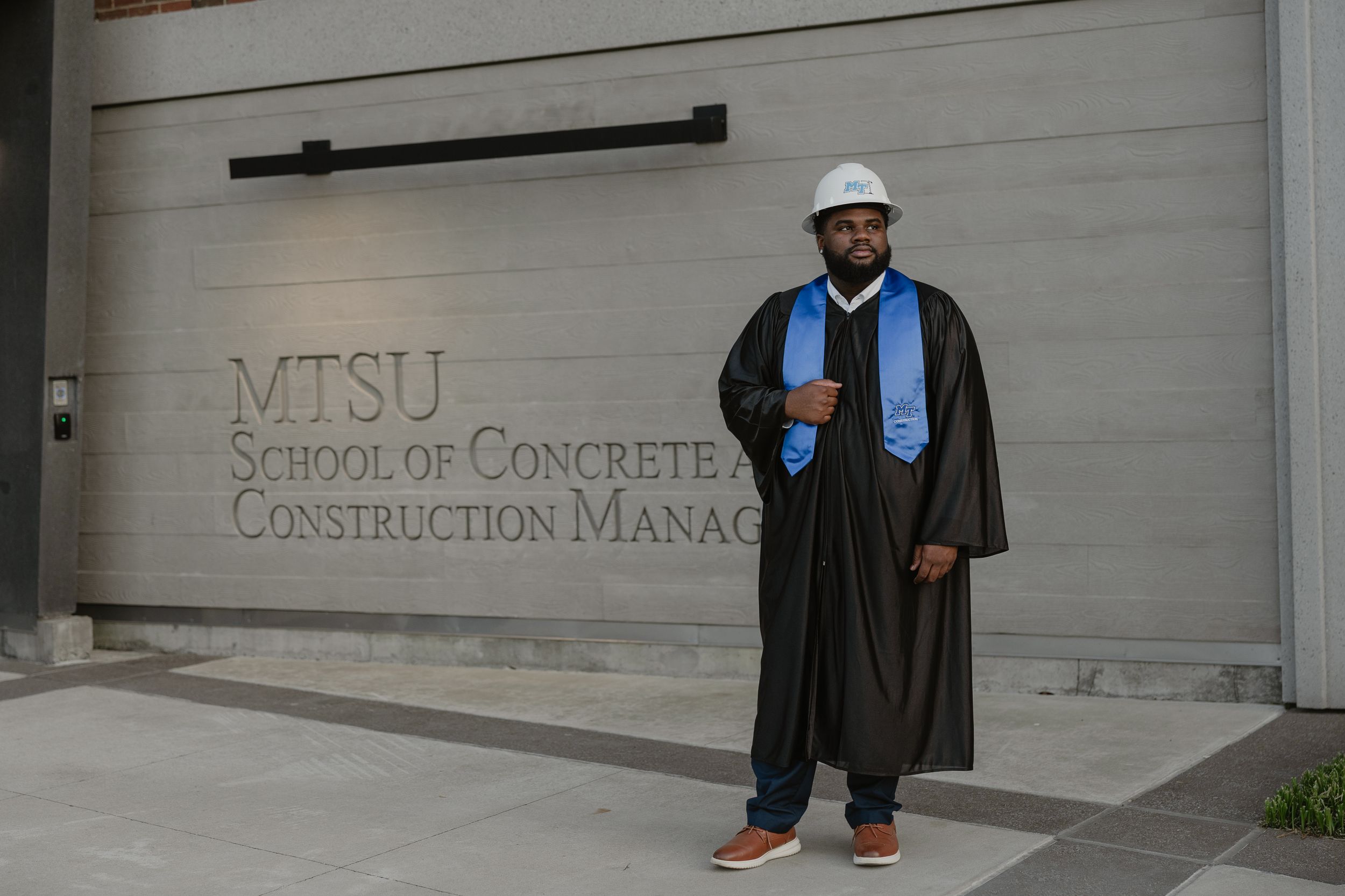 MTSU Construction Management graduate in cap, gown, hard hat, and blue stole poses proudly outside school building.