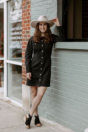 Woman in black denim dress, tan cowboy hat, and black wedge sandals posing against a mint green brick wall.