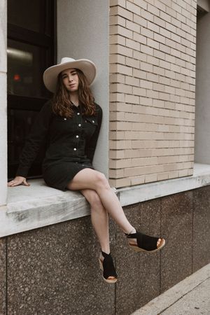 Young woman in black dress and wide-brim hat sitting on a ledge outside a brick building wearing black wedge sandals.