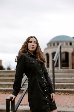 Young woman in black denim dress standing outdoors near steps with a domed building in the background.