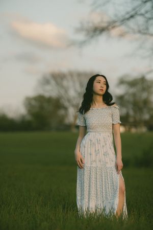 Young woman in a floral white maxi dress standing in a green field at dusk with trees in the background.