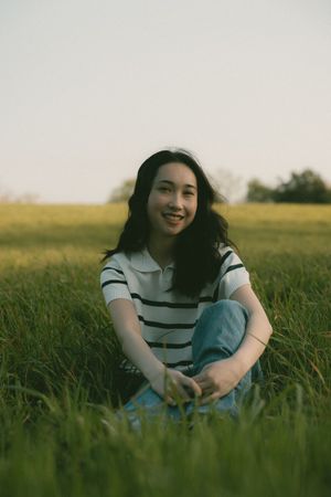 Young woman sitting cross-legged in a lush green field, smiling, wearing a striped shirt and jeans at sunset.