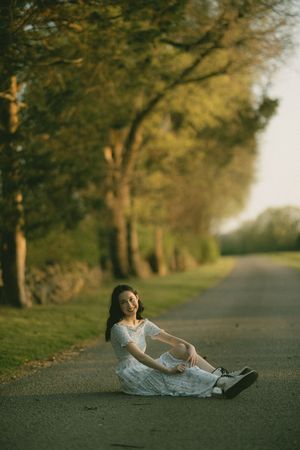Young woman in white dress sitting on a tree-lined road, smiling at camera during golden hour.