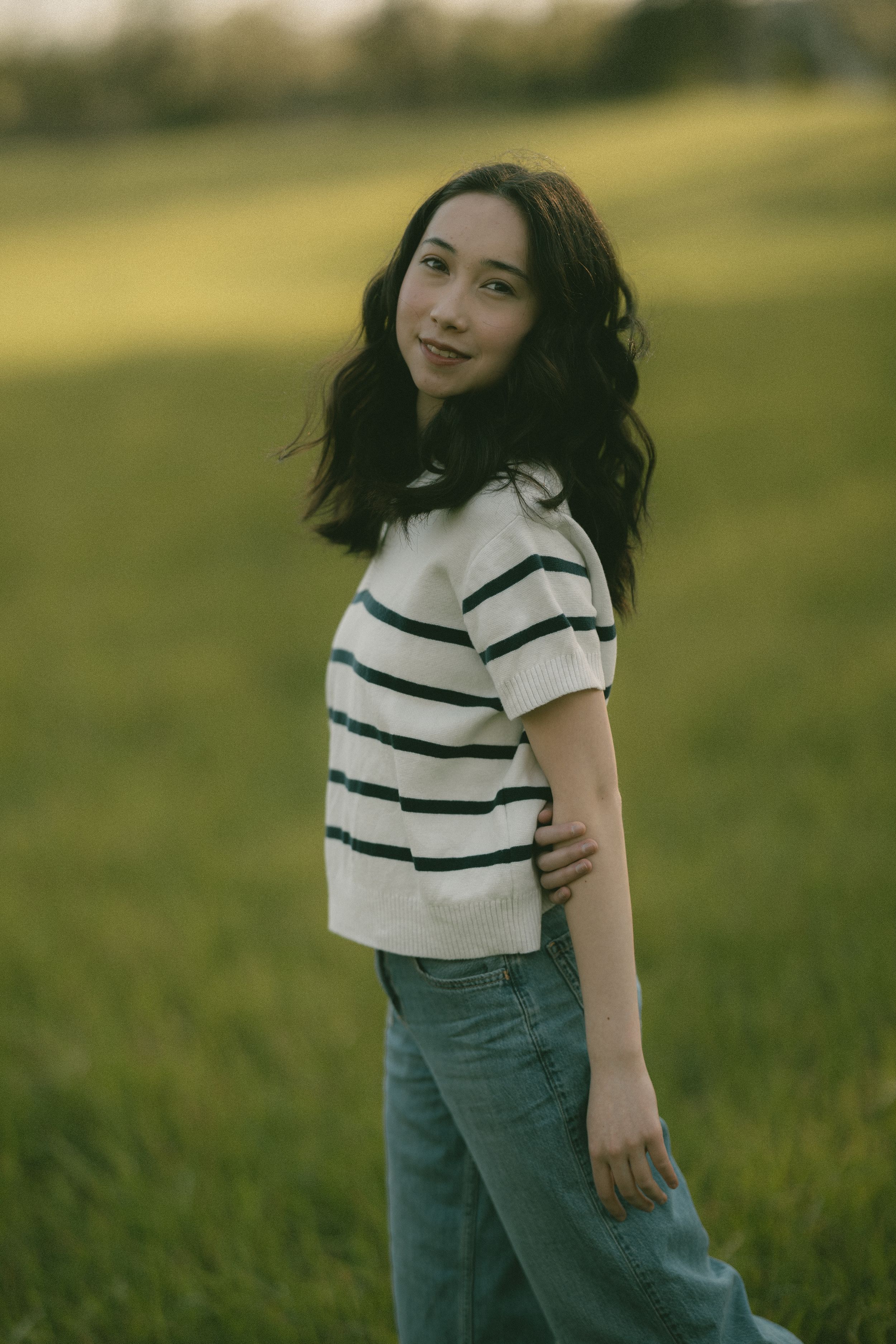 Young woman in striped crop top and jeans standing in a green field during golden hour.