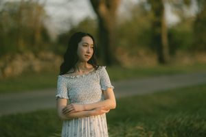Young Asian woman in floral dress standing outdoors in golden hour light, arms crossed, looking away thoughtfully.