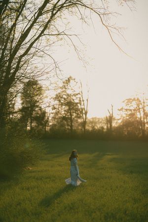 Woman in white dress standing in lush green field at golden hour sunset with bare trees in background.