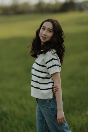 Young woman in striped white top and jeans standing in a lush green field, smiling over her shoulder.
