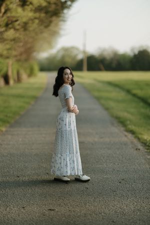 Young woman in a white floral maxi dress smiling on a tree-lined path in a serene park setting.