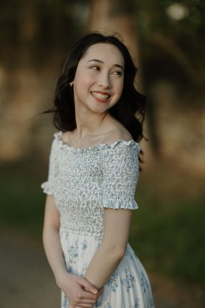 Smiling young woman in a white floral off-shoulder smocked dress posing outdoors in soft natural light.