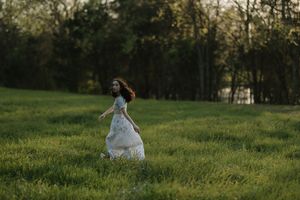 A young woman in a flowing white dress runs through a lush green meadow surrounded by trees at dusk.