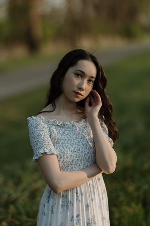 Young woman in floral smocked dress poses outdoors in golden hour light, hand raised to face.