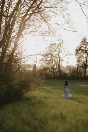 A bride in a white gown stands alone in a sunlit green meadow surrounded by trees during golden hour.