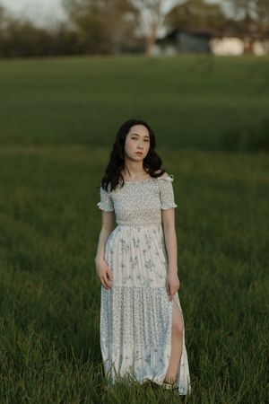 Young woman in white floral maxi dress standing in lush green field at dusk with farmhouse in background.