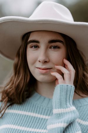 Young woman in a wide-brim white hat and light blue striped sweater, gently touching her face outdoors.