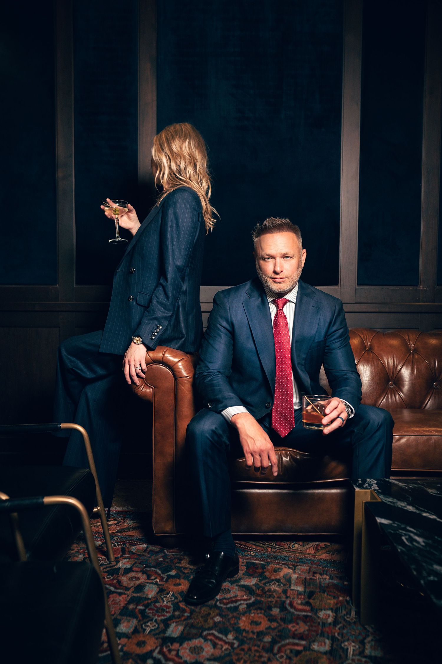 A man in a navy suit with red tie holds whiskey while a blonde woman in a suit perches on a leather sofa.