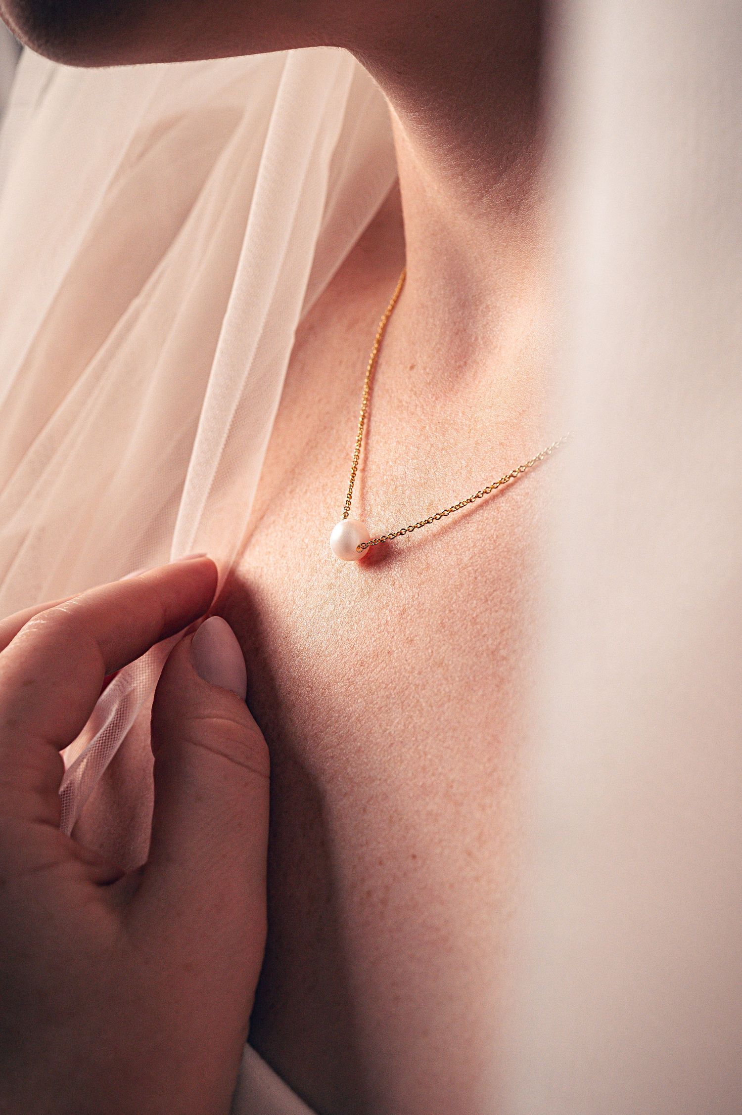 Close-up of a delicate gold pearl pendant necklace on a bride's neckline with a soft tulle veil.