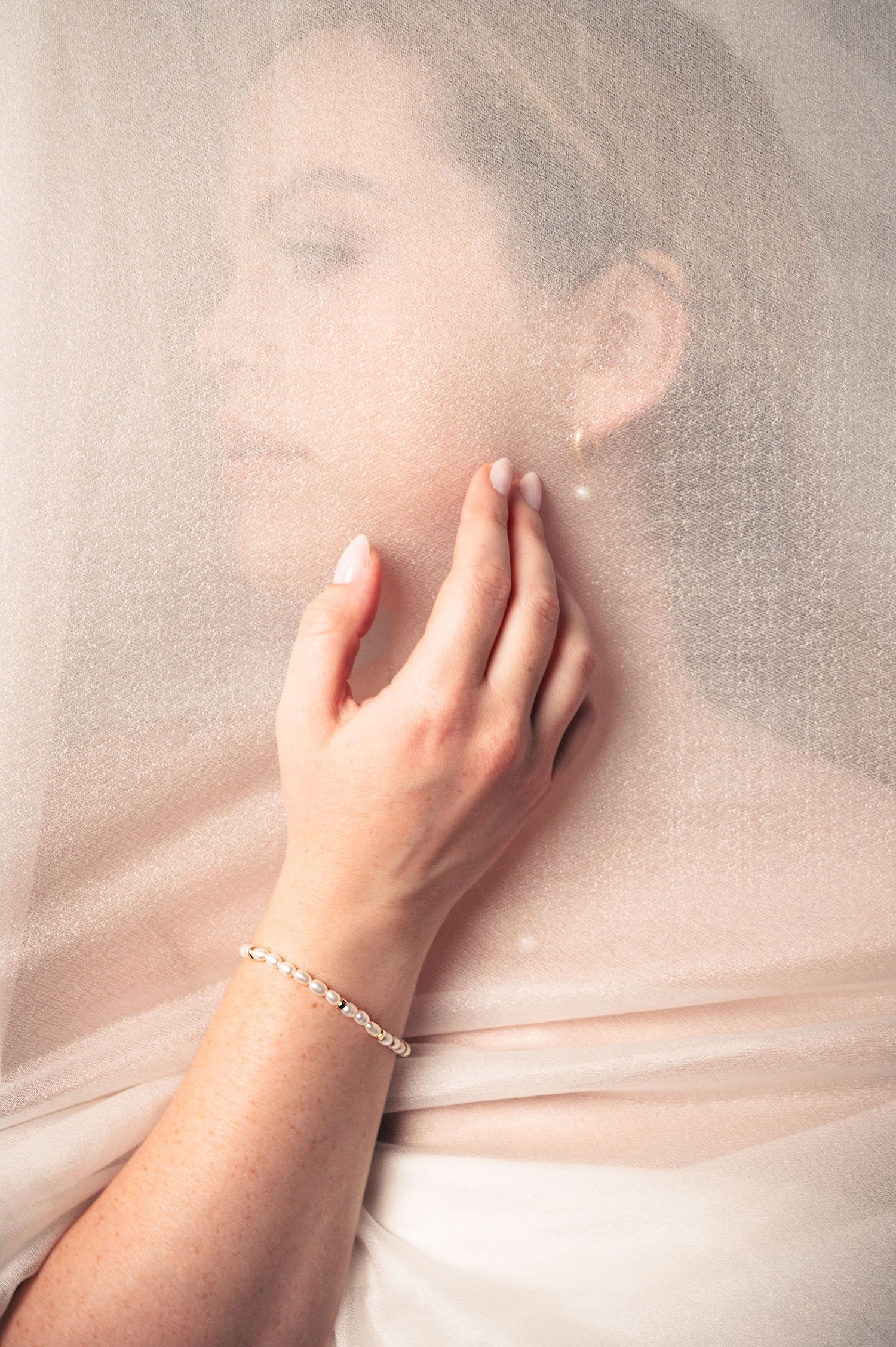 Bride in white veil touching pearl earring with delicate bracelet adorned wrist in soft romantic bridal portrait.