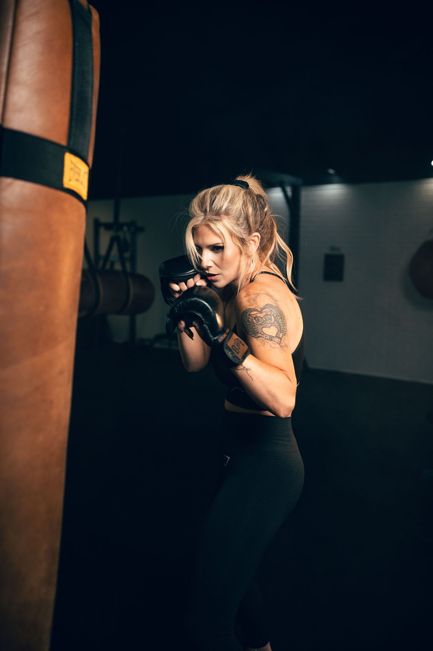 A tattooed blonde woman in boxing gloves trains with a heavy punching bag in a dark gym.