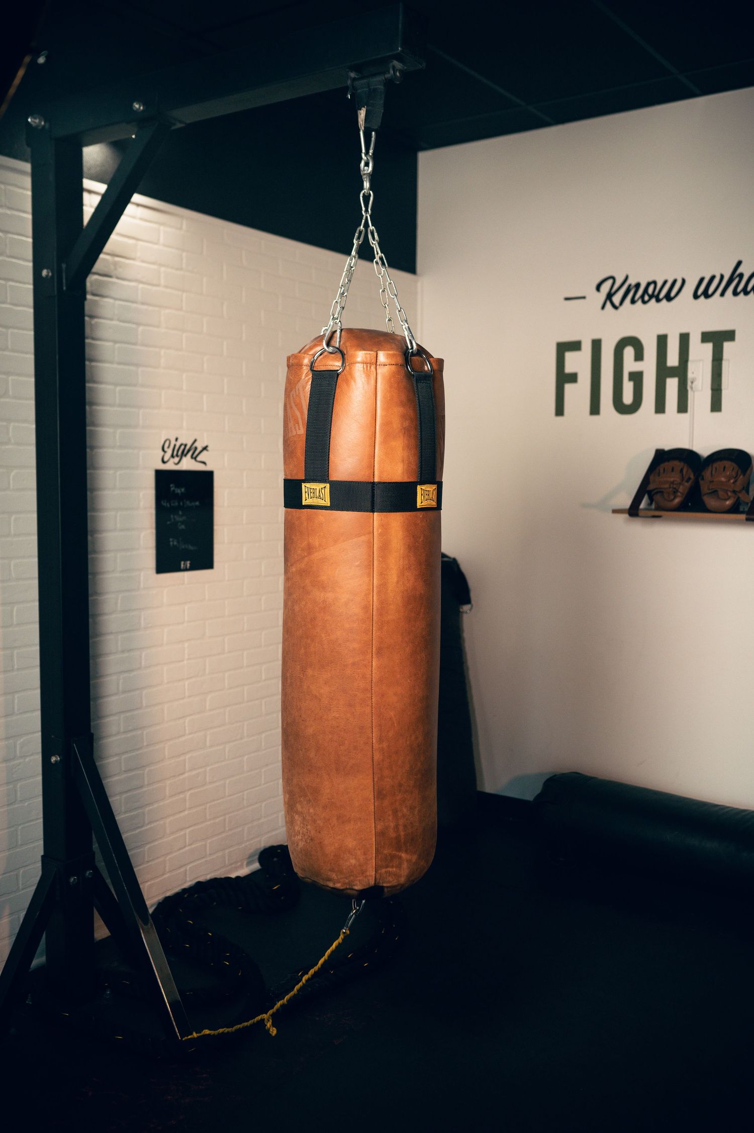 Vintage leather heavy punching bag hanging from a metal stand in a modern boxing gym with motivational wall text.