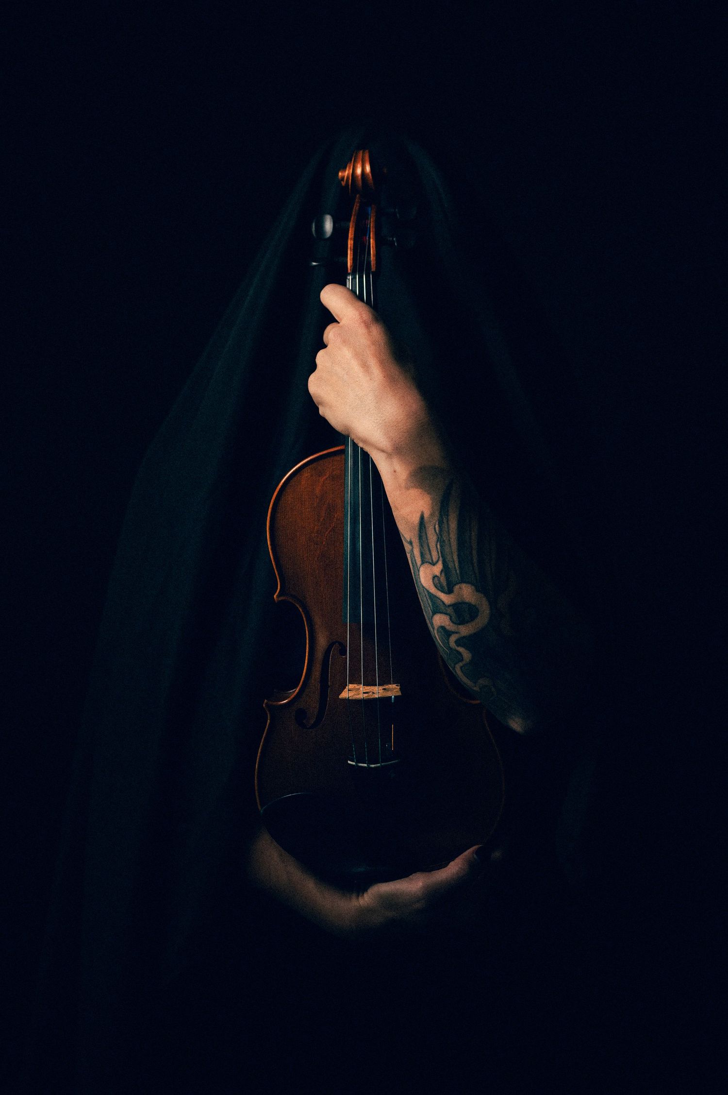 Tattooed hands hold a violin emerging from dark fabric against a black background.