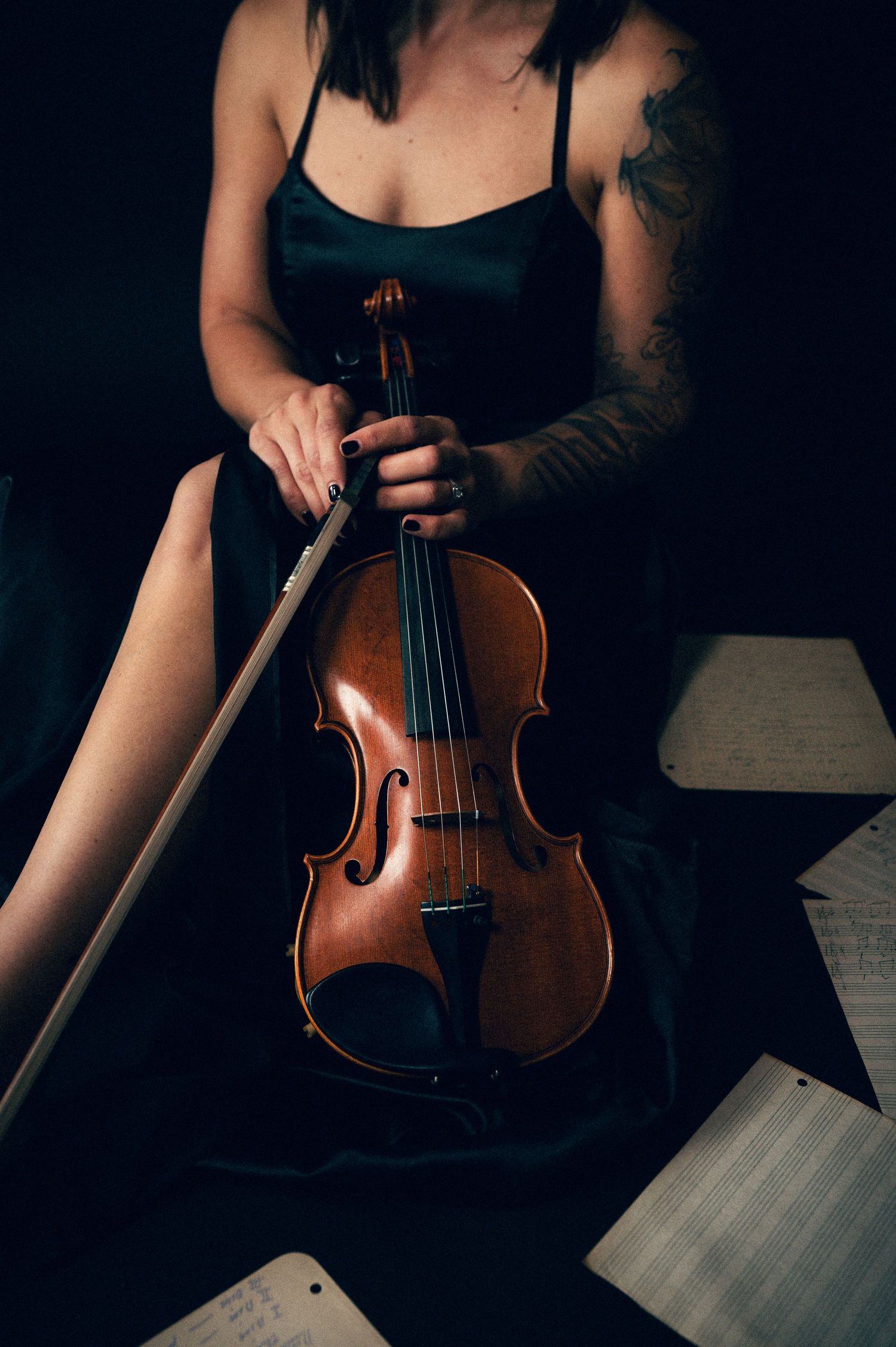 Tattooed woman in black dress holding a violin and bow with sheet music in a dark, moody setting.