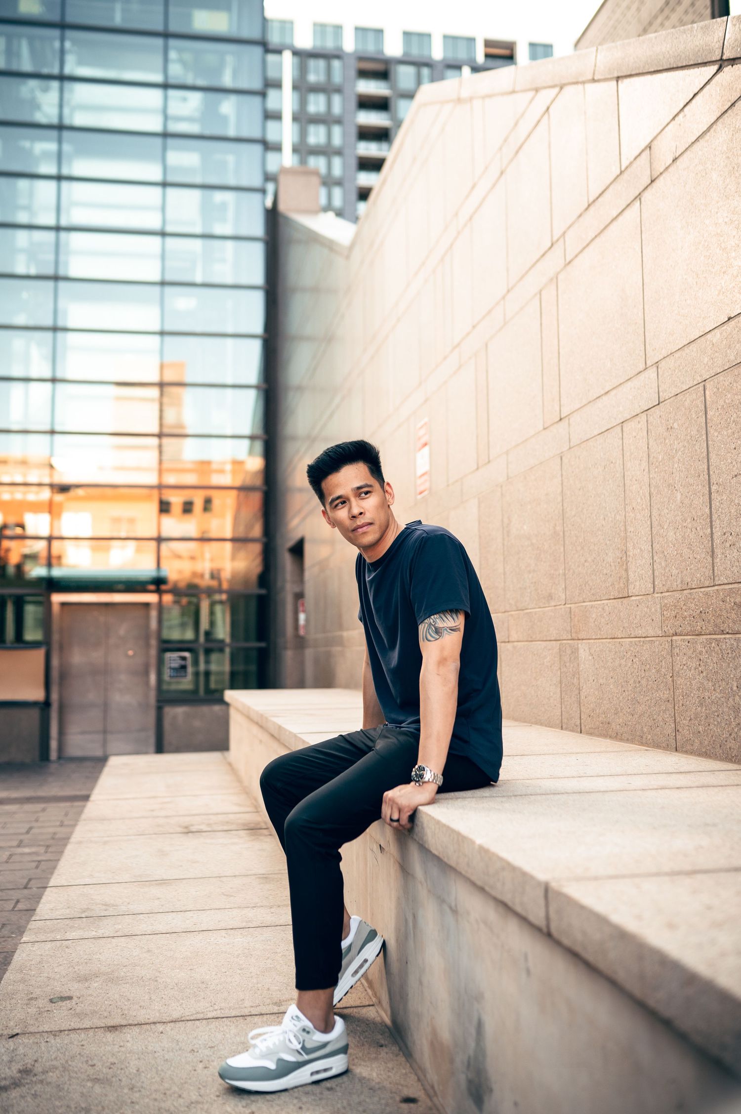 Young Asian man in navy t-shirt and black jeans sits on stone ledge in urban setting at golden hour.