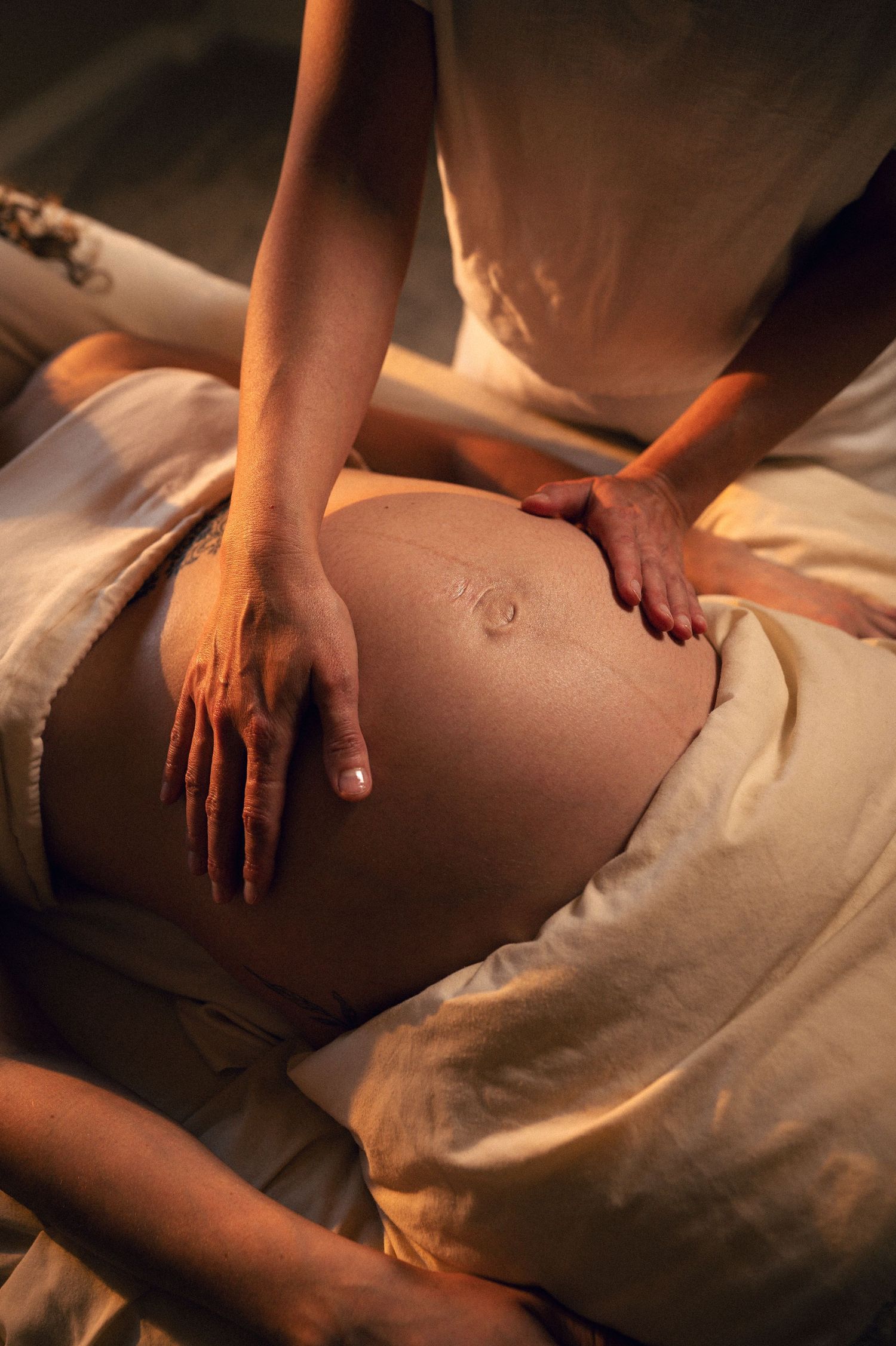 A massage therapist performing a prenatal belly massage on a pregnant woman lying on soft beige linens.