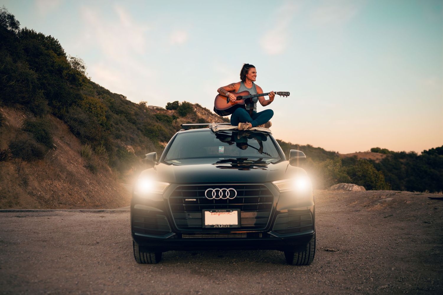 Person playing acoustic guitar while sitting on top of a black Audi SUV at dusk in a scenic outdoor setting.