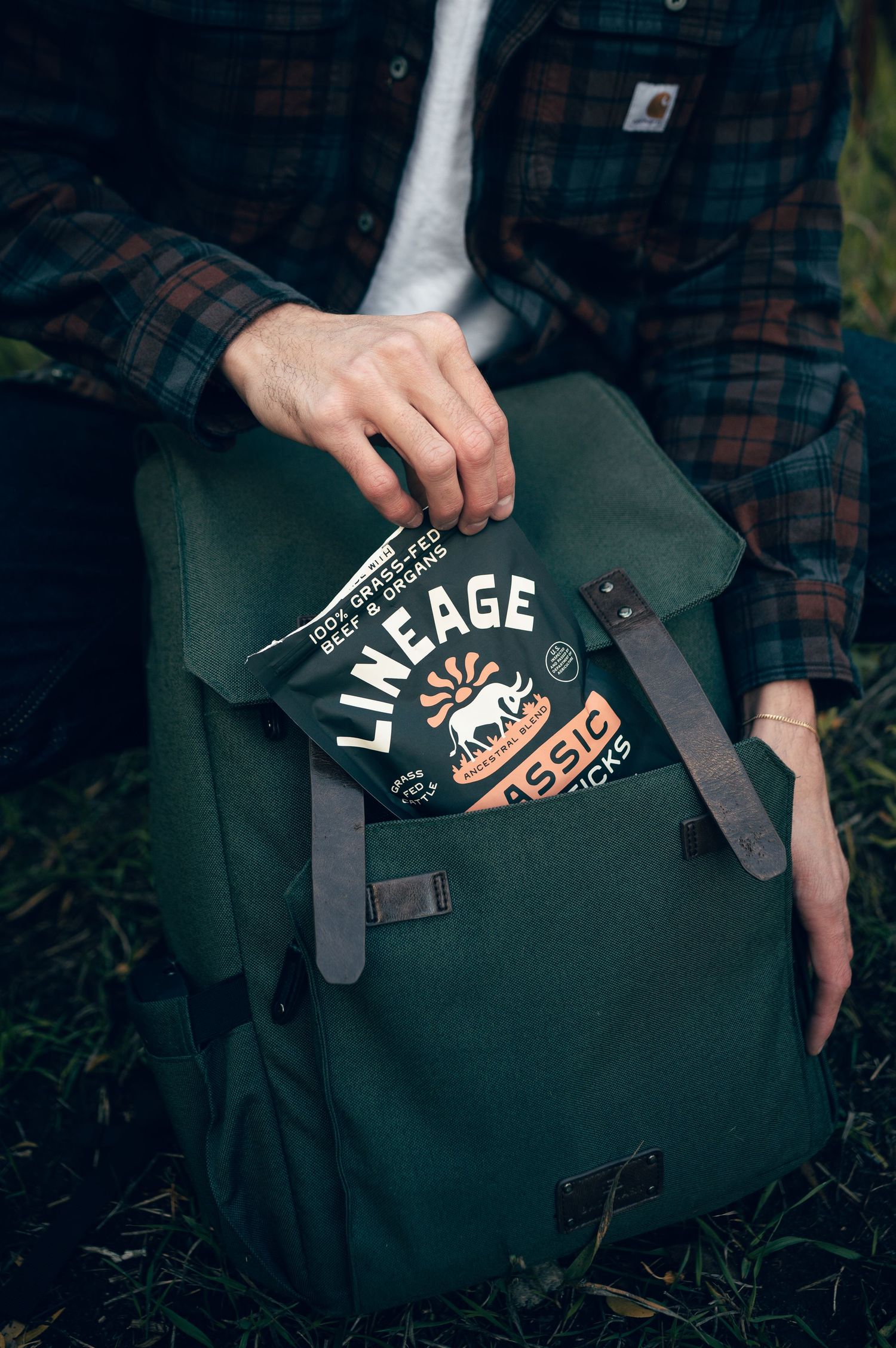 Person placing Lineage Classic meat sticks snack pack into a green canvas backpack outdoors.