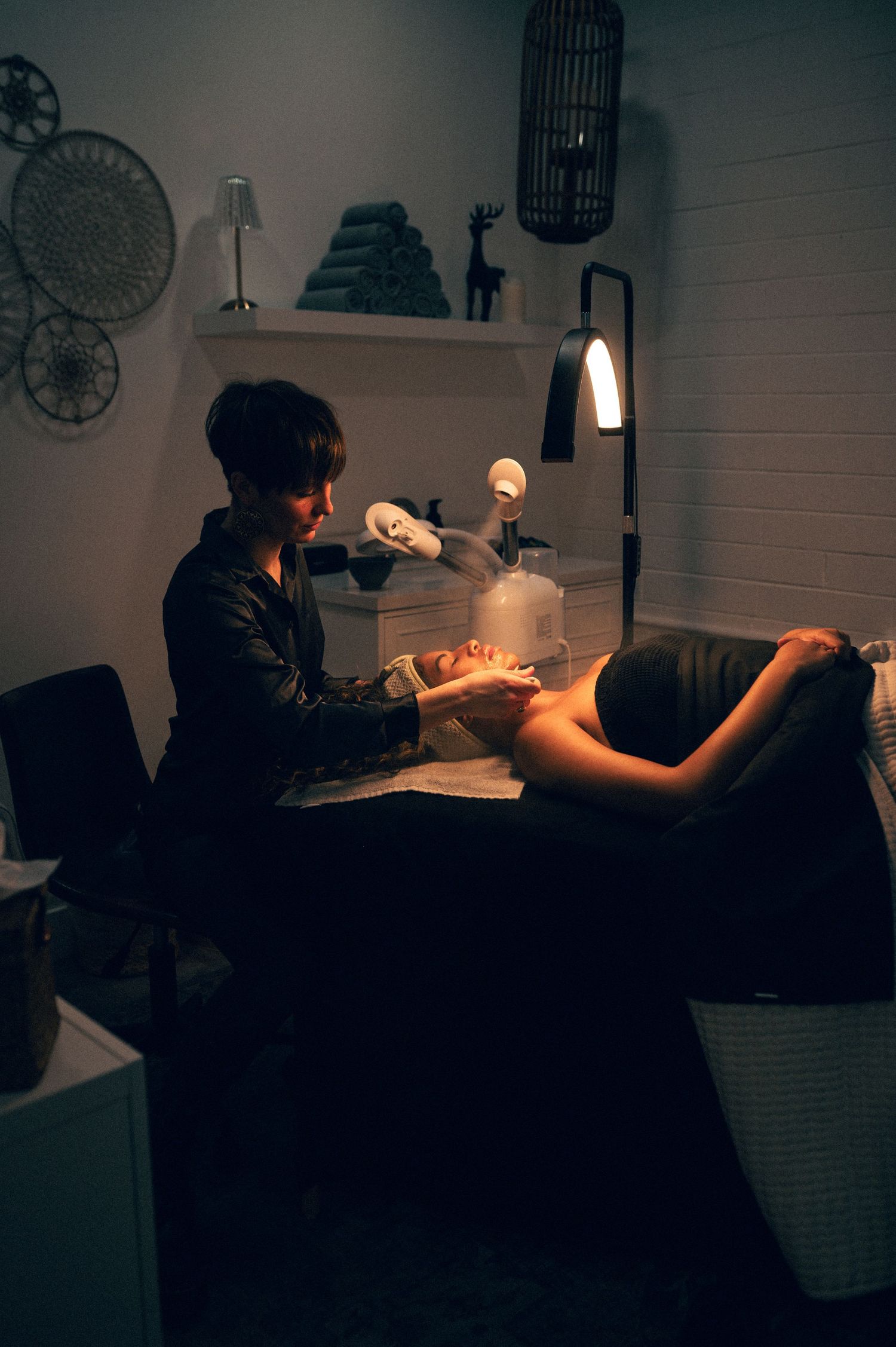 Esthetician performing a facial treatment on a client in a dimly lit spa room with decorative wall accents.