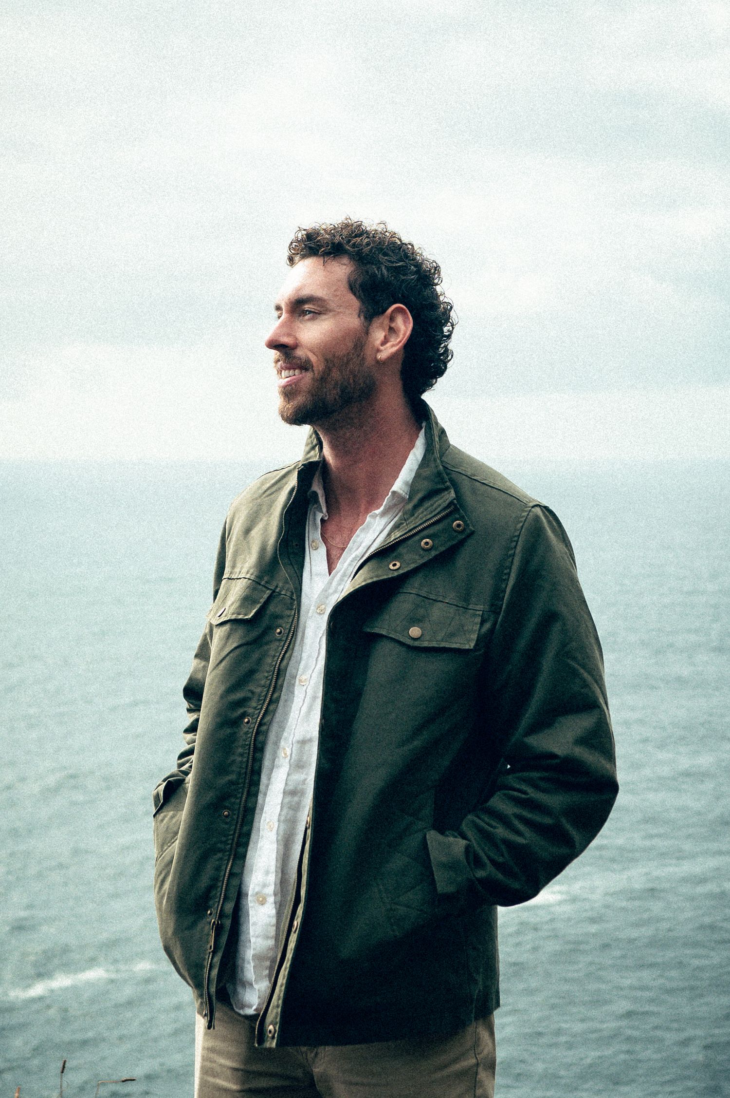 Smiling man in olive jacket stands on coastal cliff with ocean backdrop on overcast day.
