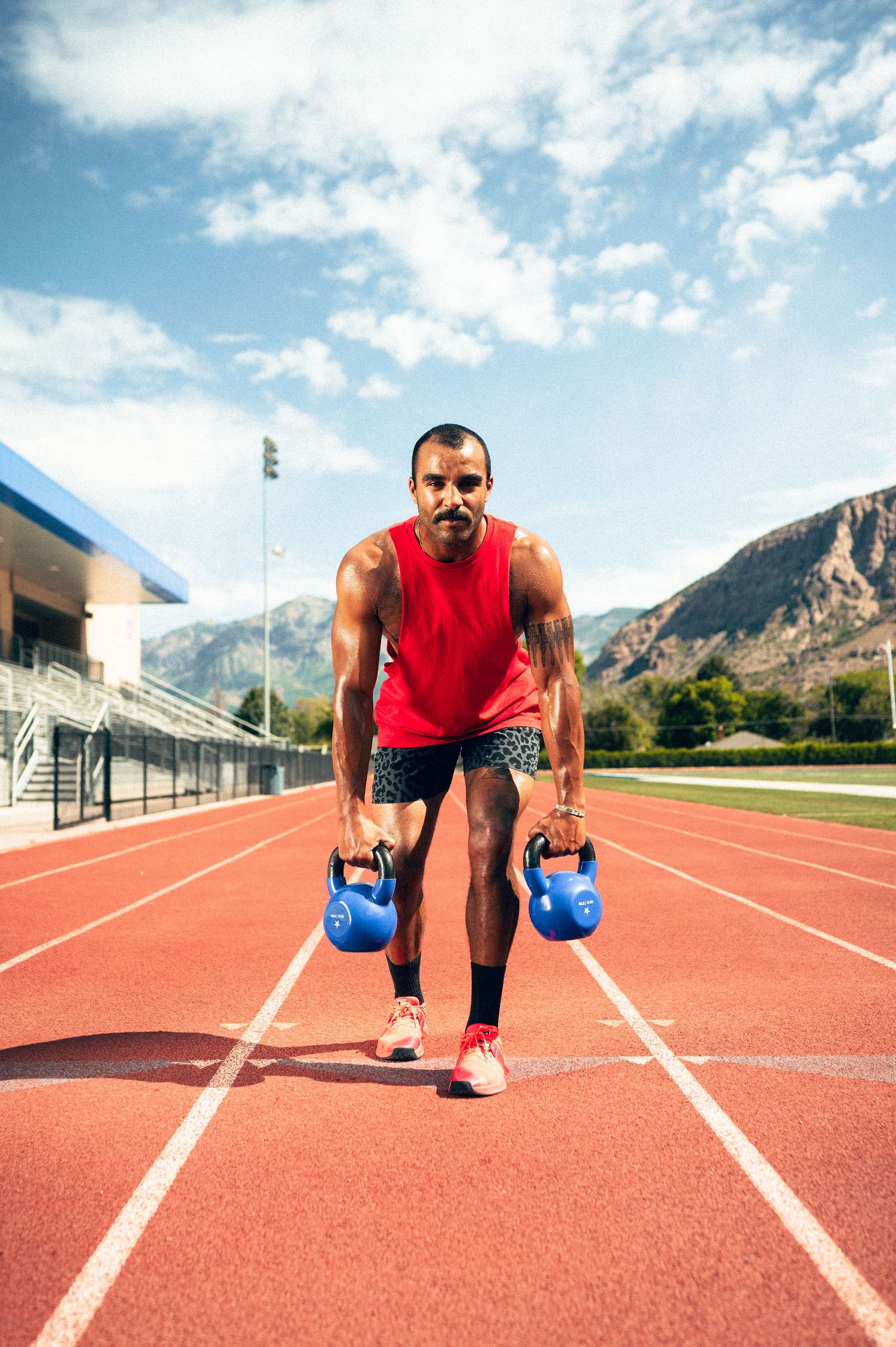 Athlete performing kettlebell exercise on red running track with mountains and blue sky in background.