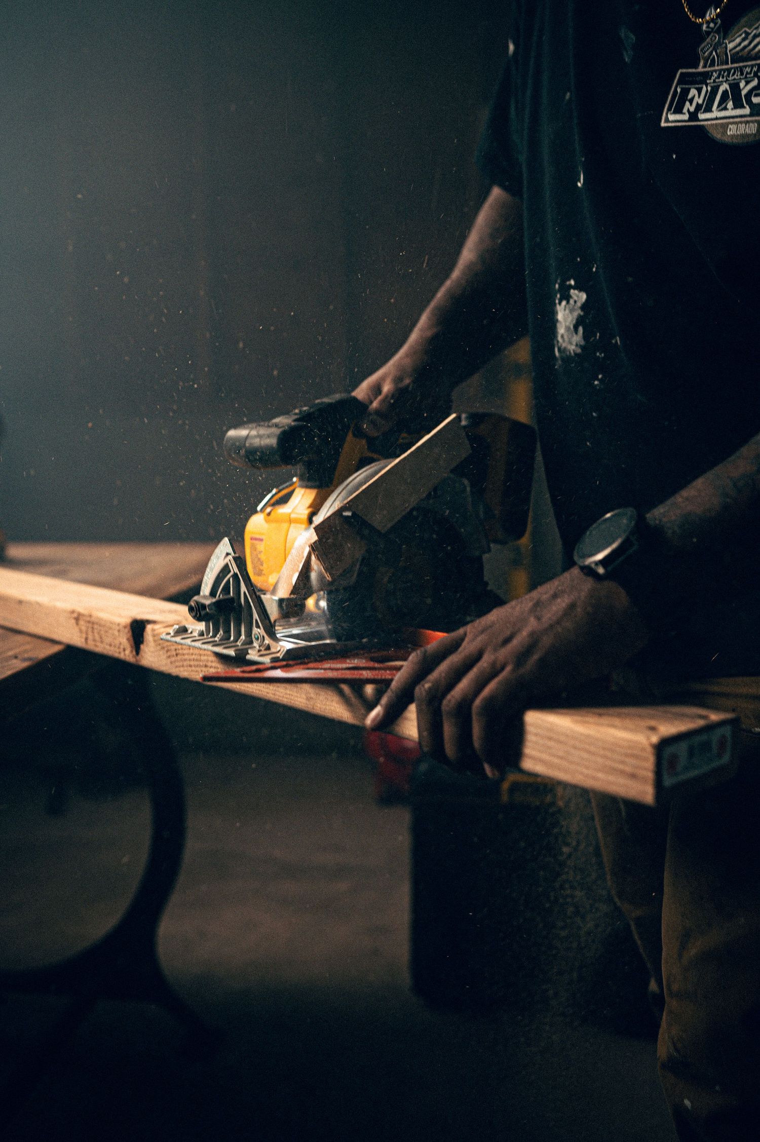 A craftsman uses a power planer on a wooden board, sending sawdust flying in a dimly lit workshop.