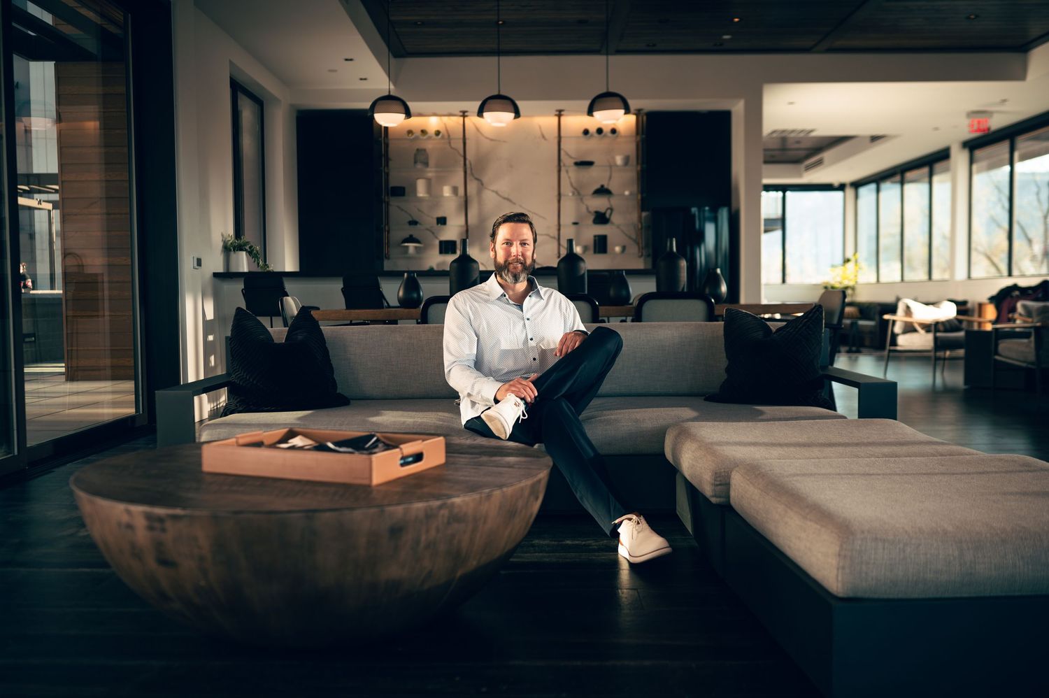 Professional man in white shirt sits confidently in modern lounge area with contemporary furniture and pendant lights.