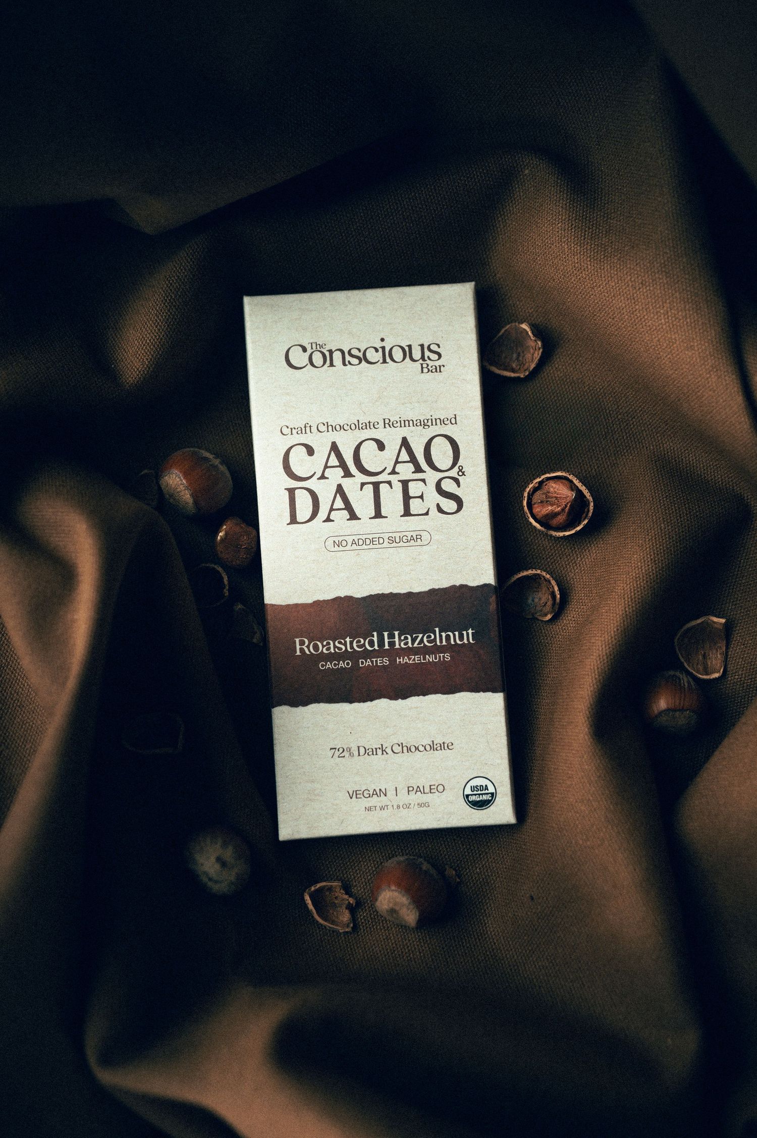 Conscious Bar Cacao Dates Roasted Hazelnut 72% dark chocolate vegan paleo bar on brown satin with hazelnuts.
