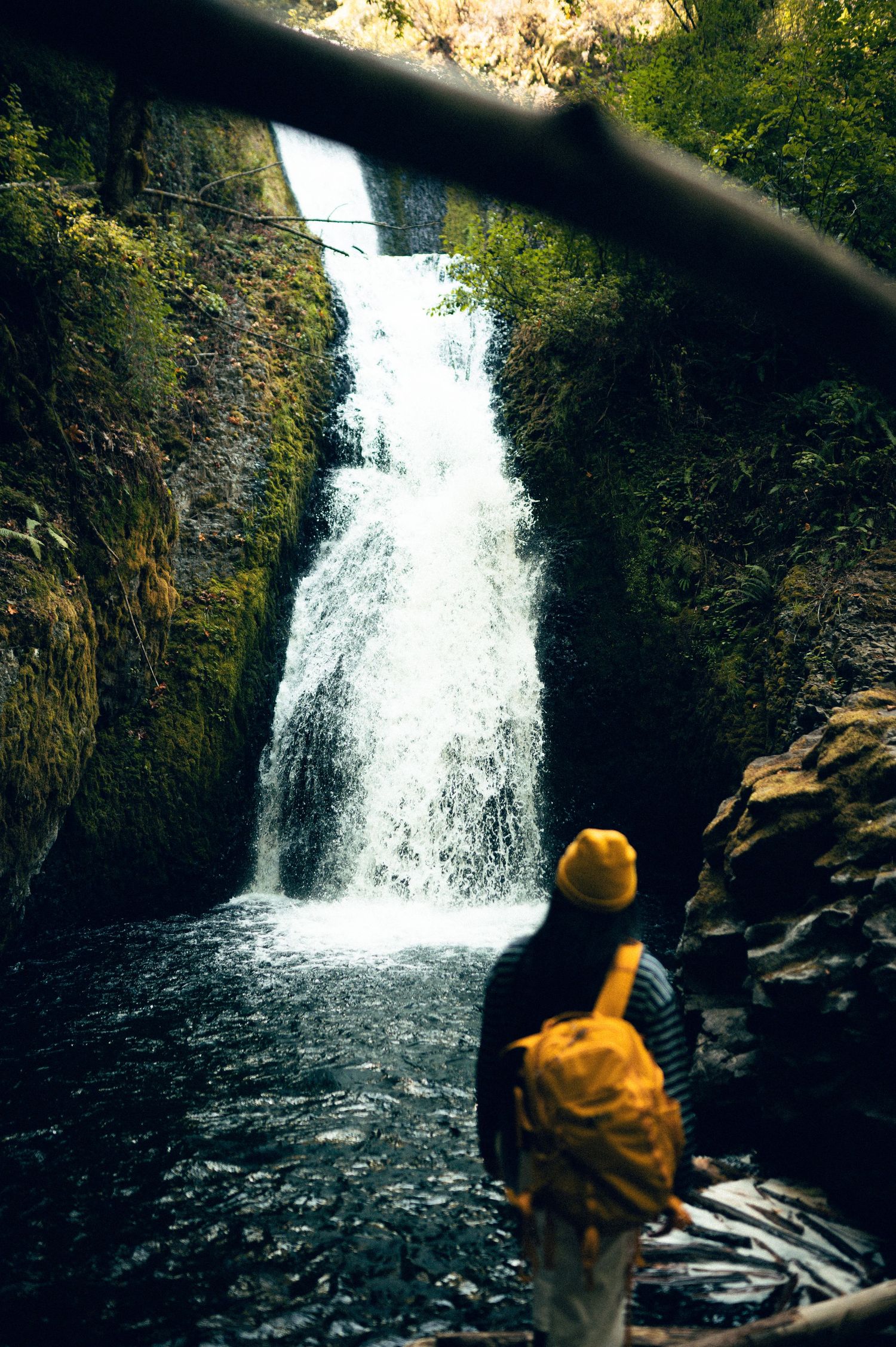 Hiker with yellow backpack and hat stands before a stunning waterfall cascading through a rocky gorge surrounded by lush greenery.