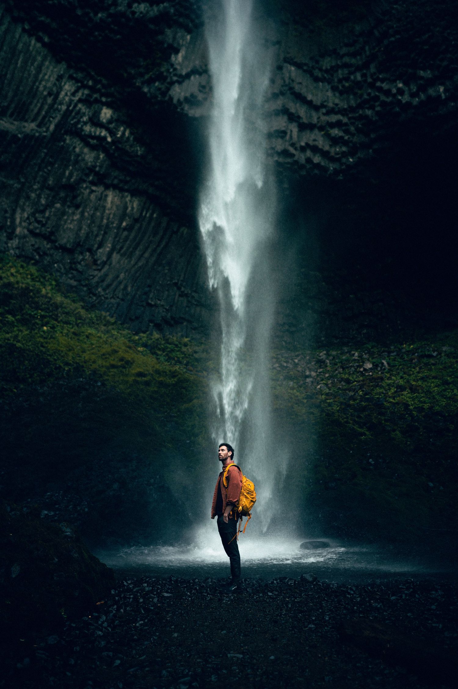 A hiker in an orange jacket stands before a towering waterfall cascading down dark rocky cliffs.