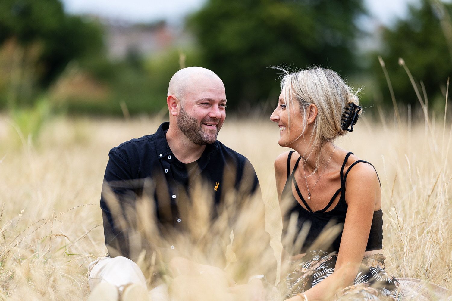 Couple sitting together smiling in a golden wheat field, enjoying a romantic outdoor moment.
