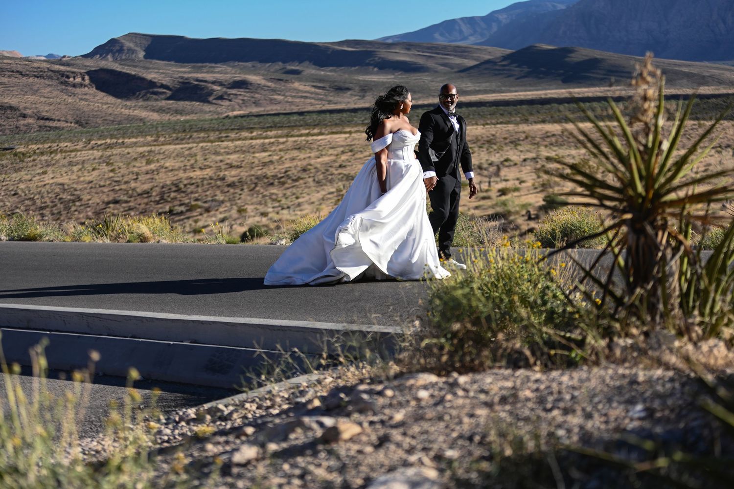 Bride and groom walk hand in hand on a desert road with mountain backdrop in their wedding attire.