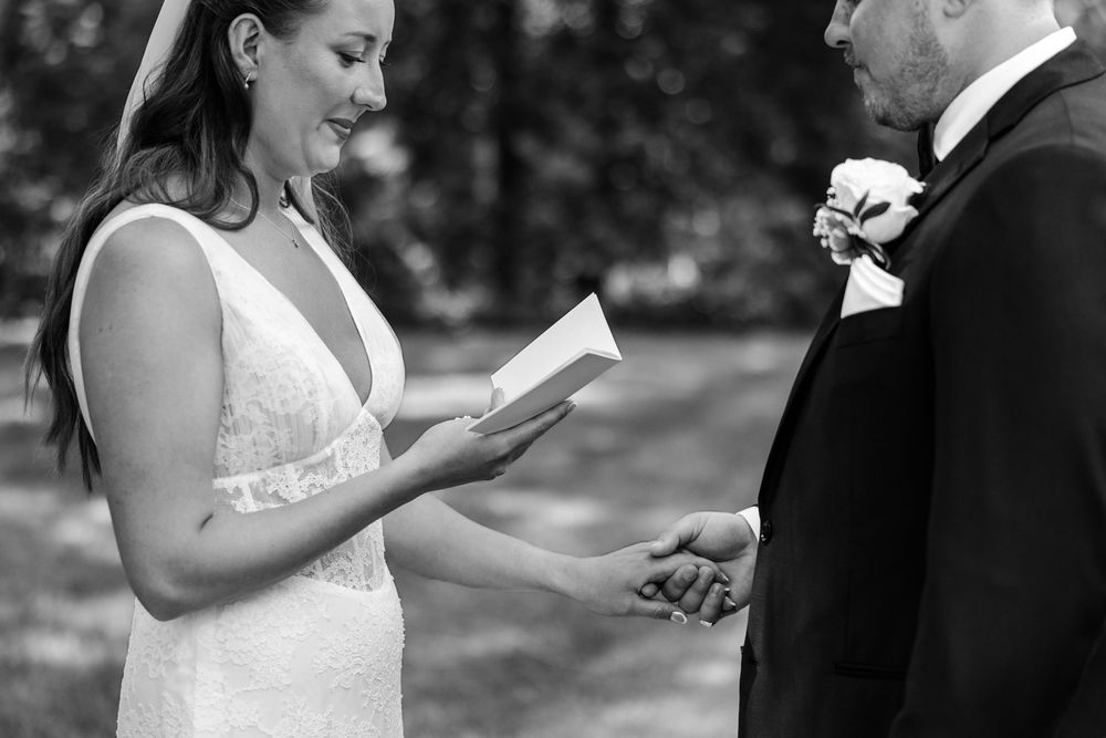 Black and white photo of a bride reading vows from a card while holding groom's hand during outdoor wedding ceremony.