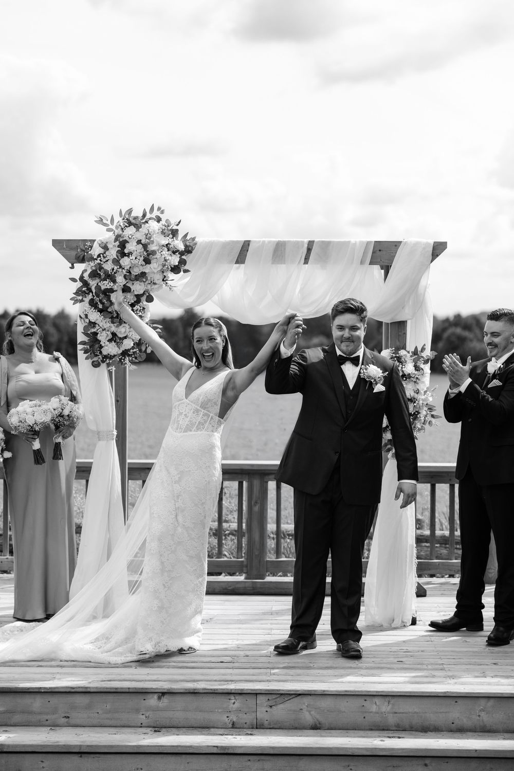 Black and white photo of a joyful bride and groom raising hands at outdoor waterfront wedding ceremony.