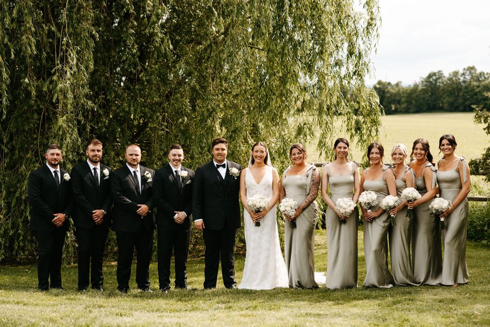 Wedding party poses under a weeping willow tree, groomsmen in black suits and bridesmaids in champagne gowns.